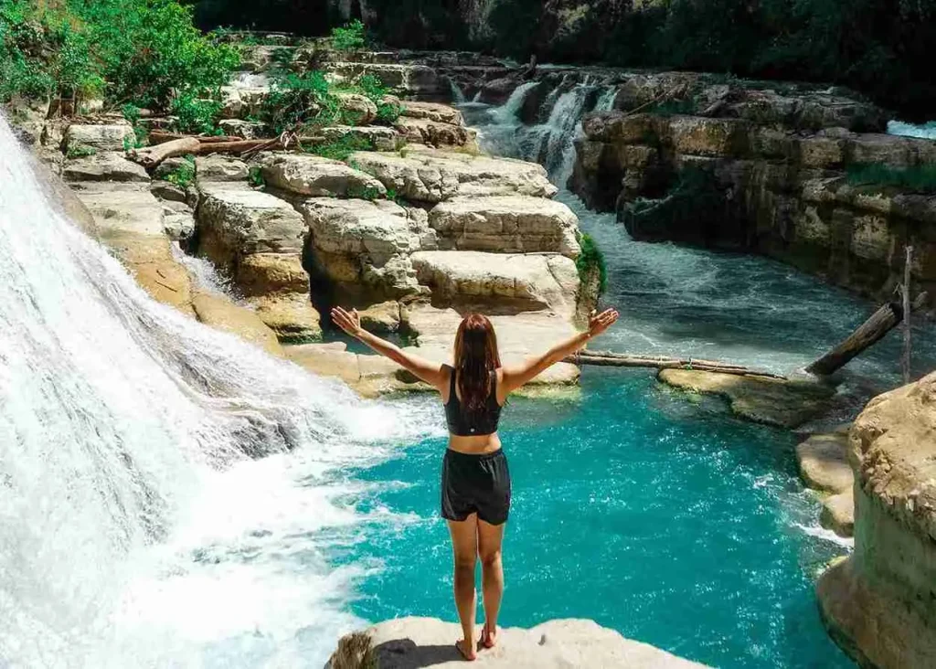 A traveller raises her arms beside the turquoise cascades of Tanggedu Waterfall in East Sumba