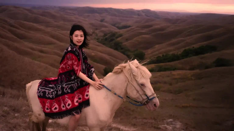 A woman in a woven ikat shawl on a white Sumba horse above the golden Wairinding hills at dusk
